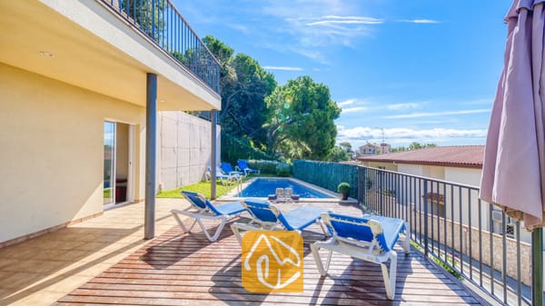 Vakantievilla's Costa Brava Spanje - Villa Mauri - Zonnig houten terras bij Villa Mauri met blauwe ligstoelen, een zwembad beneden en een overdekt terras tegen een helderblauwe hemel