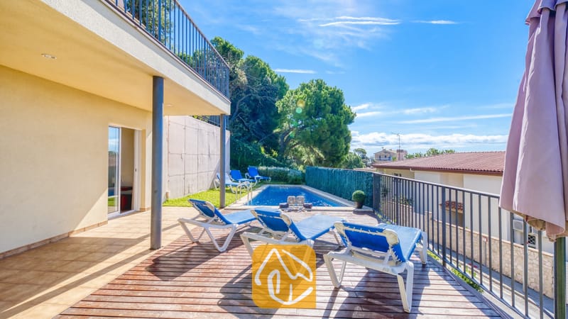 Vakantievilla's Costa Brava Spanje - Villa Mauri - Zonnig houten terras bij Villa Mauri met blauwe ligstoelen, een zwembad beneden en een overdekt terras tegen een helderblauwe hemel