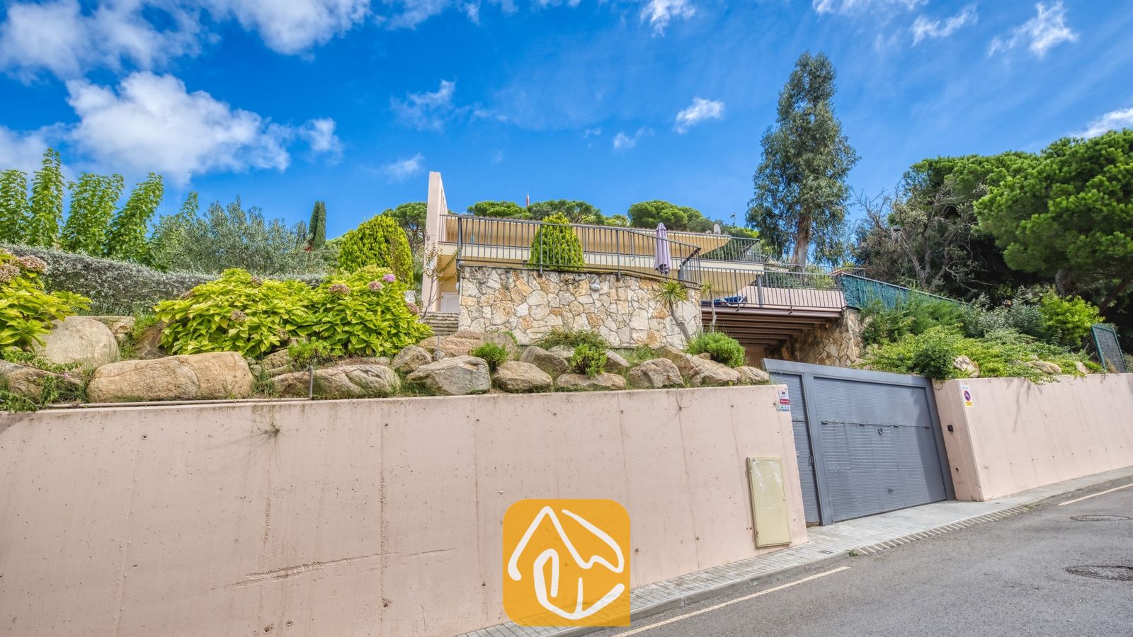 Vakantievilla's Costa Brava Spanje - Villa Mauri - Buitengevel van Villa Mauri met een stenen sokkel, een bovenbalkon met metalen leuningen en weelderige groene bomen eromheen onder een blauwe hemel.