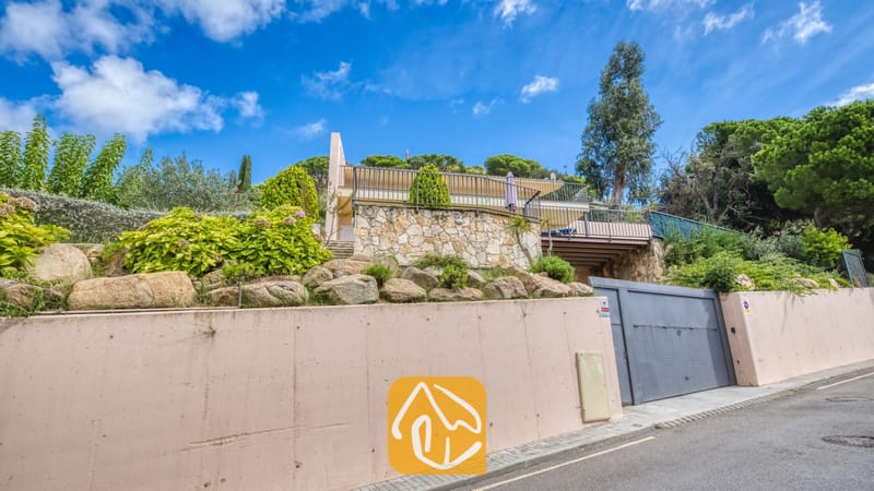 Vakantievilla's Costa Brava Spanje - Villa Mauri - Buitengevel van Villa Mauri met een stenen sokkel, een bovenbalkon met metalen leuningen en weelderige groene bomen eromheen onder een blauwe hemel.