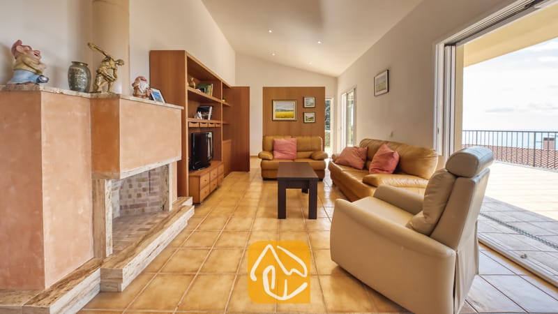 Vakantievilla's Costa Brava Spanje - Villa Mauri - Woonkamer van Villa Mauri voorzien van beige lederen banken, een grote stenen open haard en glazen schuifpuien die uitkomen op een balkon.