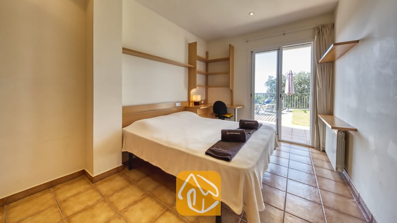 Vakantievilla's Costa Brava Spanje - Villa Mauri - Slaapkamer met een tweepersoonsbed, ingebouwde houten planken en glazen schuifdeuren die uitkomen op een zonnig buitenterras.