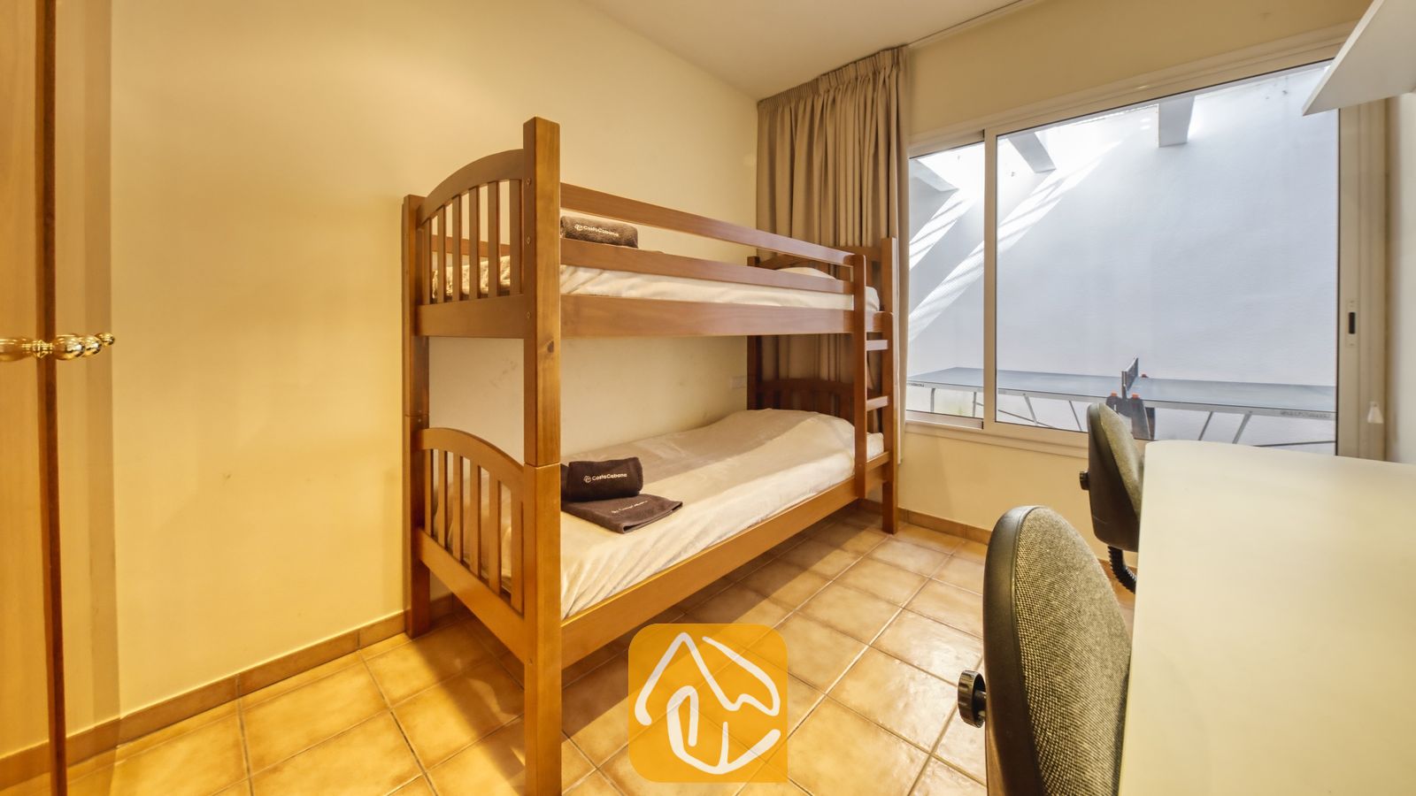 Vakantievilla's Costa Brava Spanje - Villa Mauri - Slaapkamer voorzien van een houten stapelbed met opgevouwen donkere handdoeken, tegelvloeren, een bureau met bureaustoelen en een raam met gordijnen.