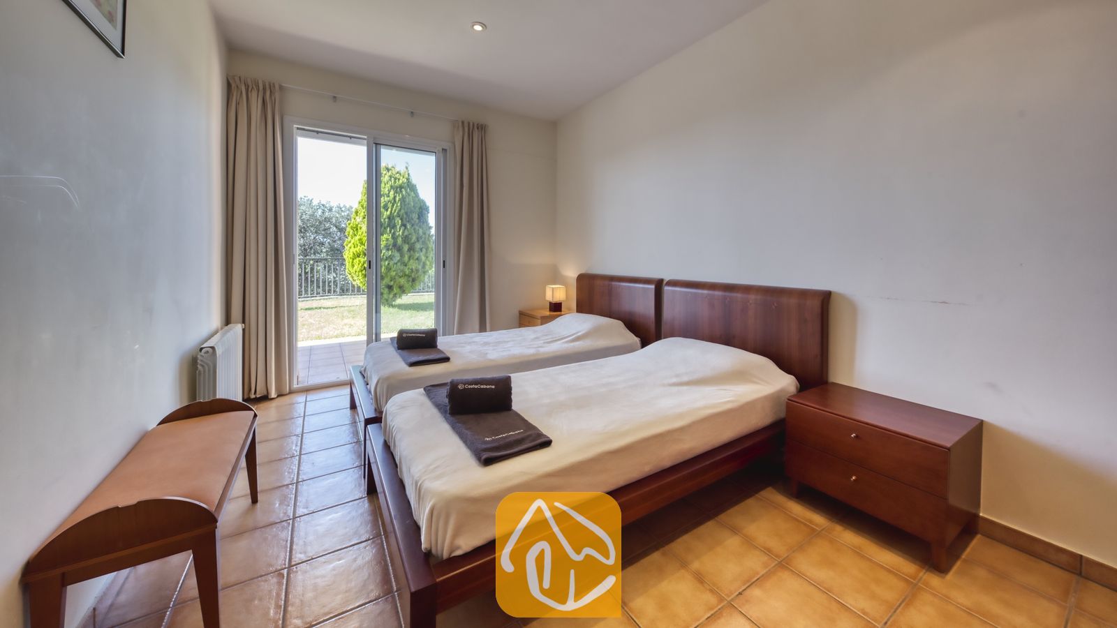 Vakantievilla's Costa Brava Spanje - Villa Mauri - Slaapkamer met twee houten eenpersoonsbedden, donker opgevouwen handdoeken, een tegelvloer en een glazen schuifpui die uitkomt op een groen buitenterras