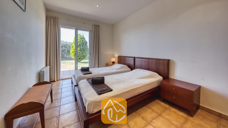 Vakantievilla's Costa Brava Spanje - Villa Mauri - Slaapkamer met twee houten eenpersoonsbedden, donker opgevouwen handdoeken, een tegelvloer en een glazen schuifpui die uitkomt op een groen buitenterras