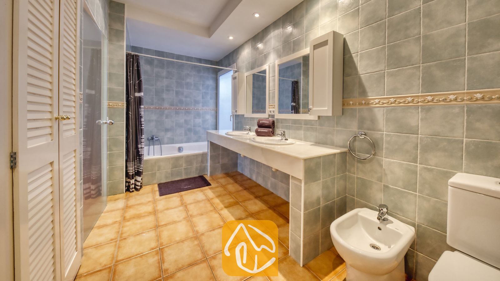 Vakantievilla's Costa Brava Spanje - Villa Mauri - Badkamer met lichtblauwe wandtegels, twee witte wastafels met bijpassende spiegels, een ligbad met een donker gordijn en terracotta vloertegels.