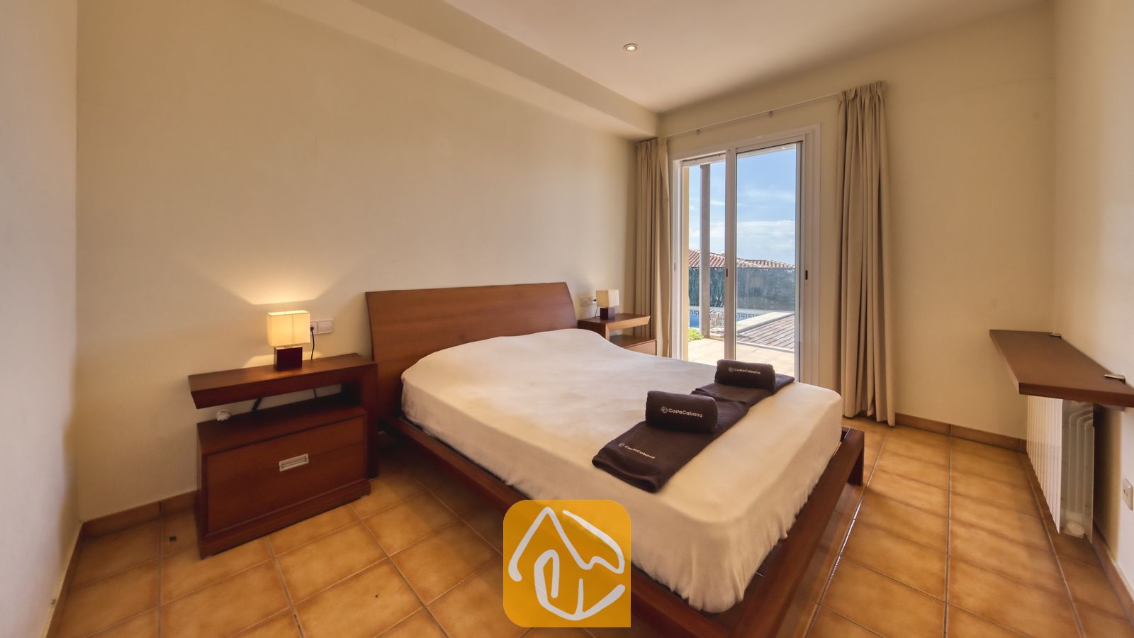 Vakantievilla's Costa Brava Spanje - Villa Mauri - Slaapkamer met houten bedframe, tegelvloer, nachtkastje met lamp en een glazen schuifpui die toegang biedt tot een terras.