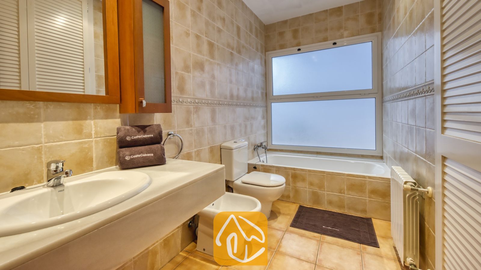 Vakantievilla's Costa Brava Spanje - Villa Mauri - Badkamer met beige betegelde muren, een witte wastafel met een houten kast erboven, een bidet, een toilet en een ligbad onder een matglasraam.