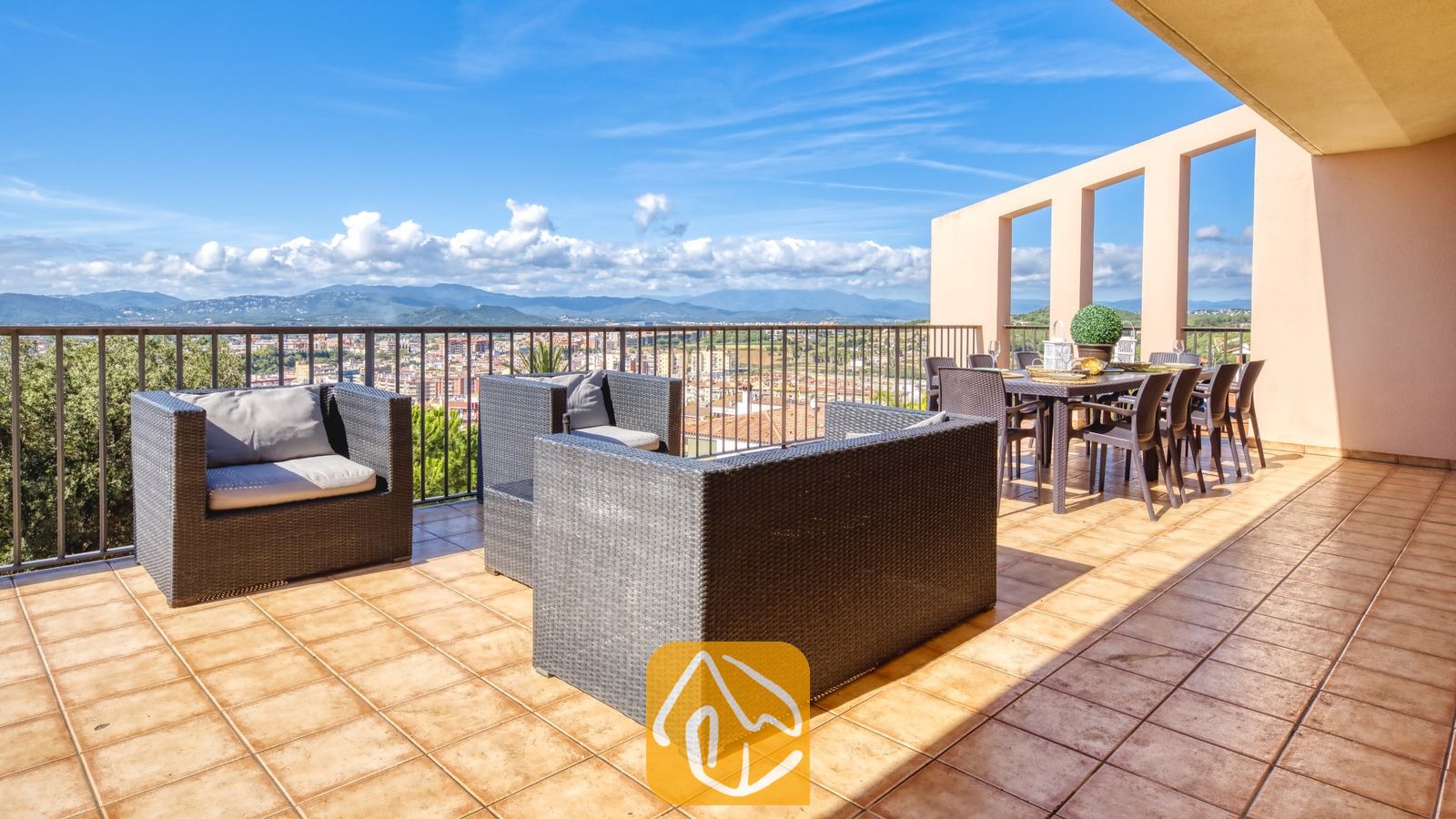 Vakantievilla's Costa Brava Spanje - Villa Mauri - Terras met uitzicht op de bergen en het dorp bij Villa Mauri, voorzien van donkerkleurige rieten fauteuils met grijze kussens en een eettafel onder een colonnade.