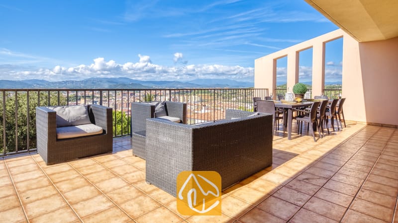 Vakantievilla's Costa Brava Spanje - Villa Mauri - Terras met uitzicht op de bergen en het dorp bij Villa Mauri, voorzien van donkerkleurige rieten fauteuils met grijze kussens en een eettafel onder een colonnade.