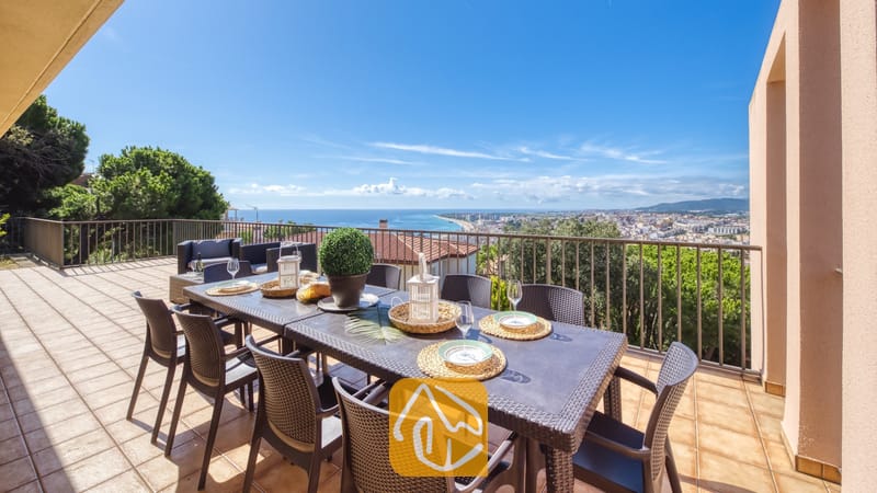 Vakantievilla's Costa Brava Spanje - Villa Mauri - Terras van Villa Mauri met een grote, donkere wicker eettafel voorzien van geweven placemats, witte lantaarns en wijnglazen, tegen een achtergrond van kust- en zeezichten.