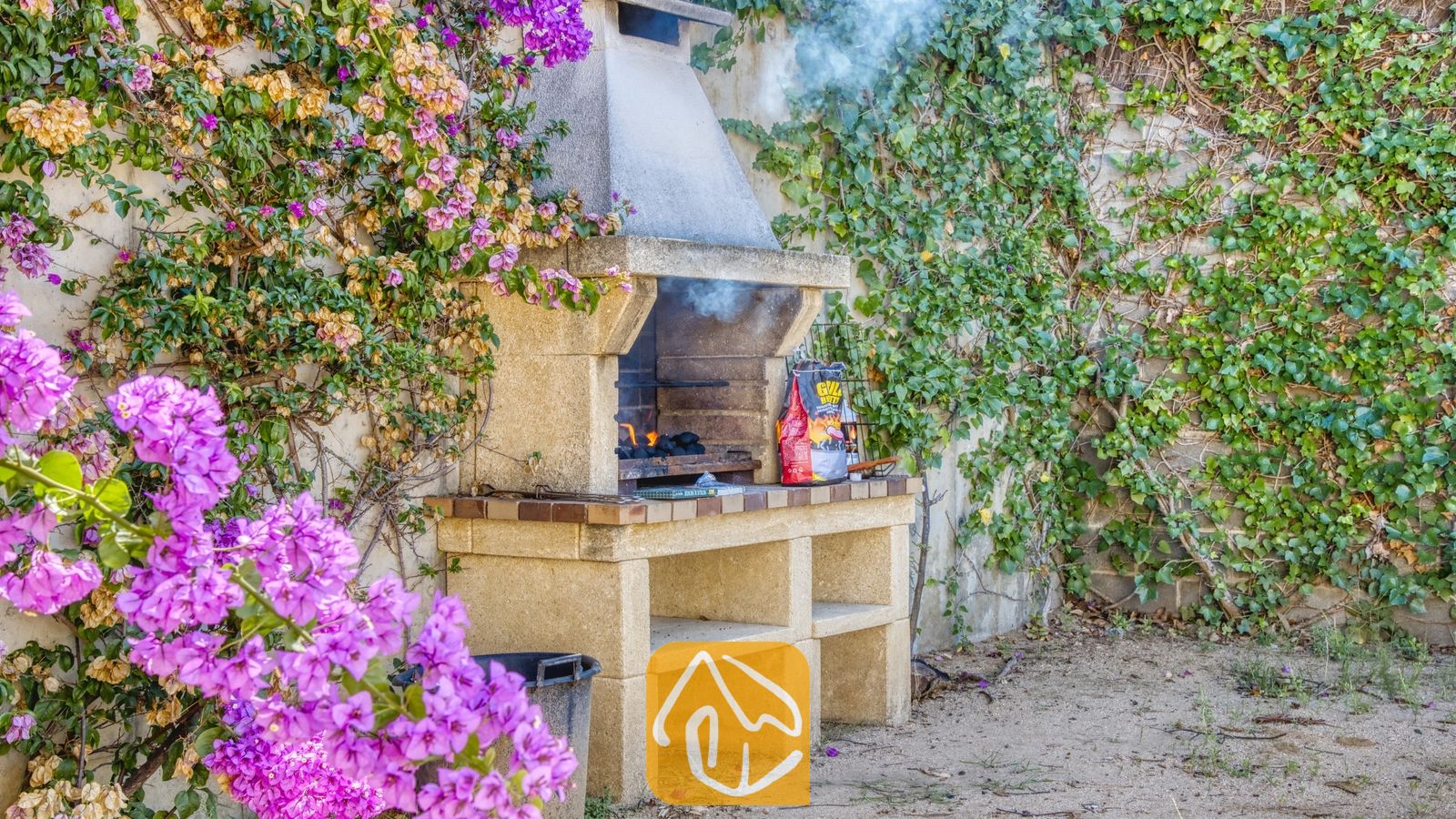 Vakantievilla's Costa Brava Spanje - Villa Mauri - Stenen buitenbarbecue bij Villa Mauri, prachtig gelegen tegen een muur begroeid met groene klimop en felpaarse bougainville.