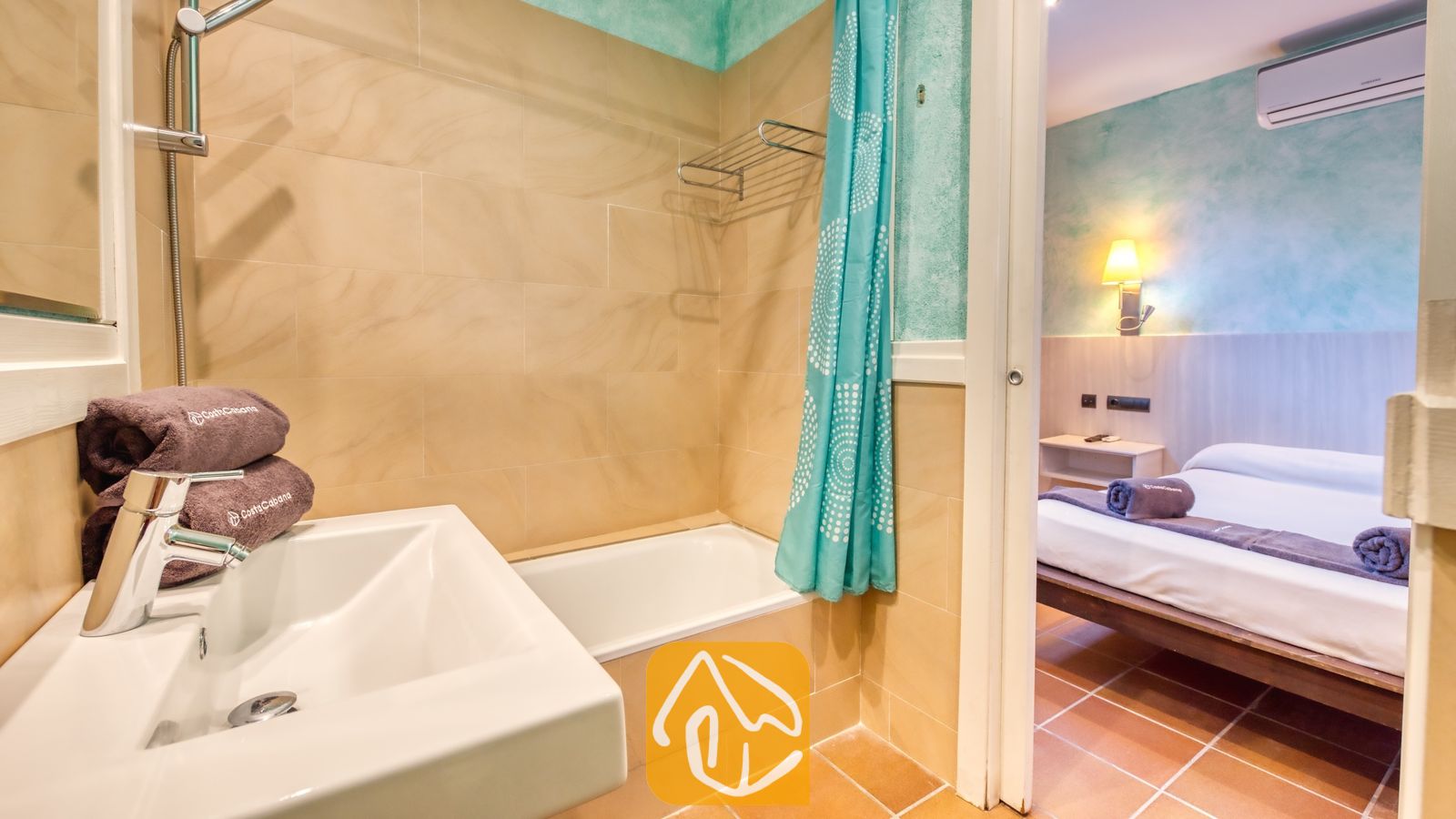 Vakantievilla's Costa Brava Countryside Spanje - Villa Can Bernardi - Badkamer met een witte keramische wastafel met opgestapelde bruine handdoeken en een ligbad met een turquoise patroongordijn, die uitkomt op een aangrenzende slaapkamer.