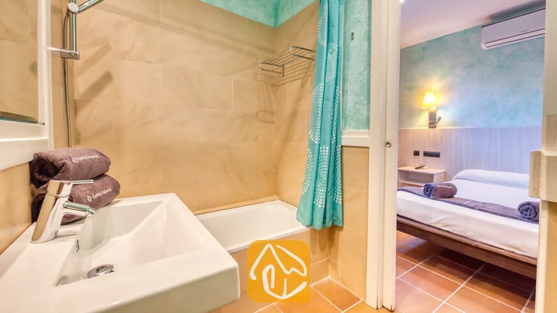 Villas de vacances Costa Brava Countryside Espagne - Villa Can Bernardi - Salle de bains comprenant un lavabo en céramique blanche surmonté de serviettes marron empilées et une baignoire ornée d'un rideau à motif turquoise, ouvrant sur une chambre attenante.