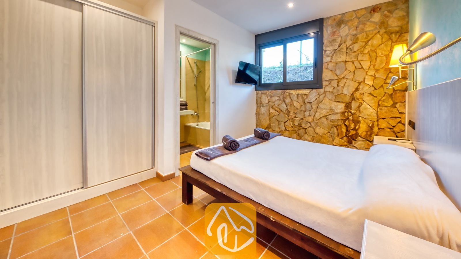 Vakantievilla's Costa Brava Countryside Spanje - Villa Can Bernardi - Slaapkamer met een tweepersoonsbed, een rustieke stenen accentmuur, een tegelvloer en een deuropening naar de en-suite badkamer.