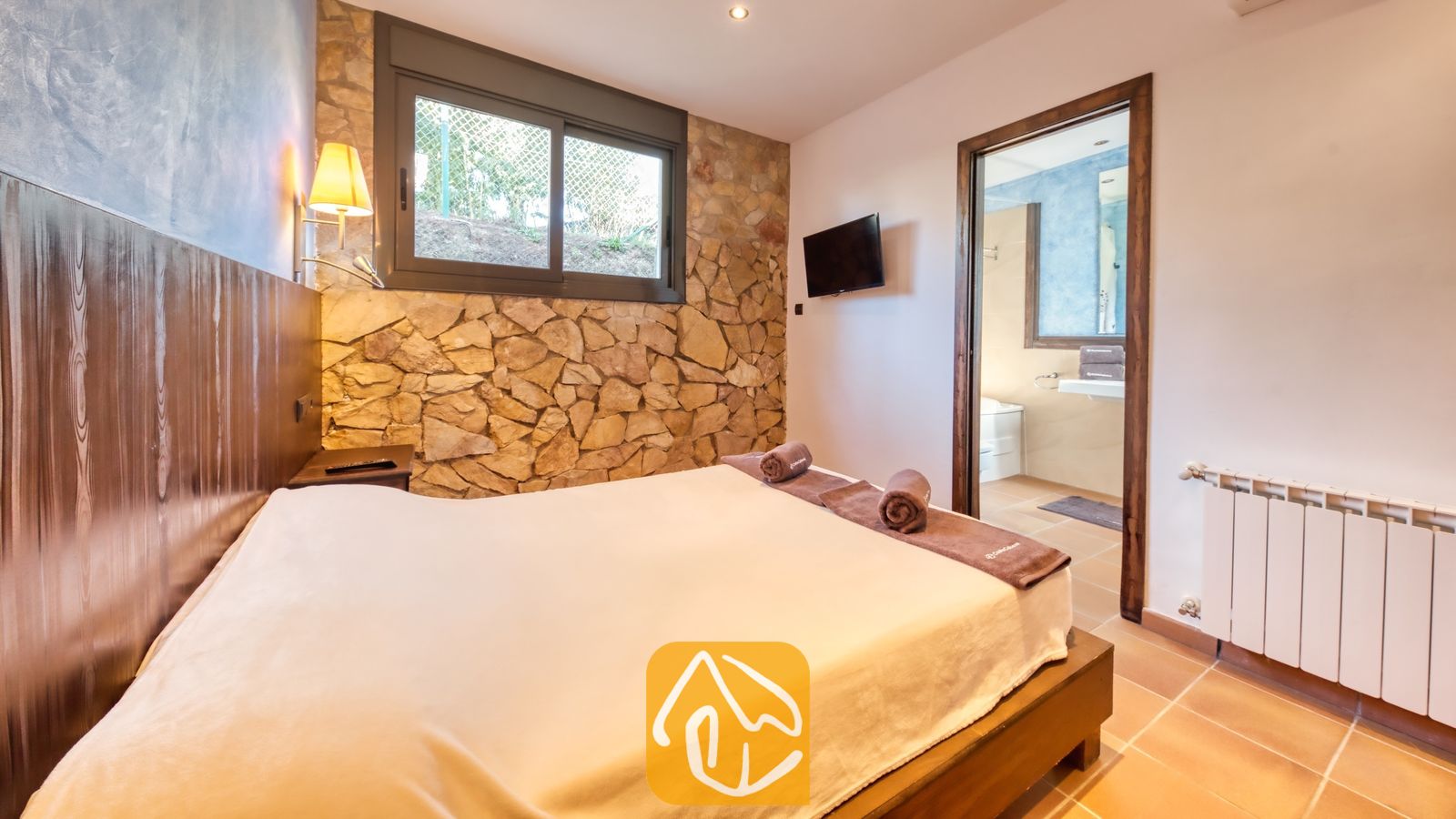 Vakantievilla's Costa Brava Countryside Spanje - Villa Can Bernardi - Slaapkamer met een tweepersoonsbed, opgerolde bruine handdoeken, een stenen accentmuur en een open deur die leidt naar een en-suite badkamer.