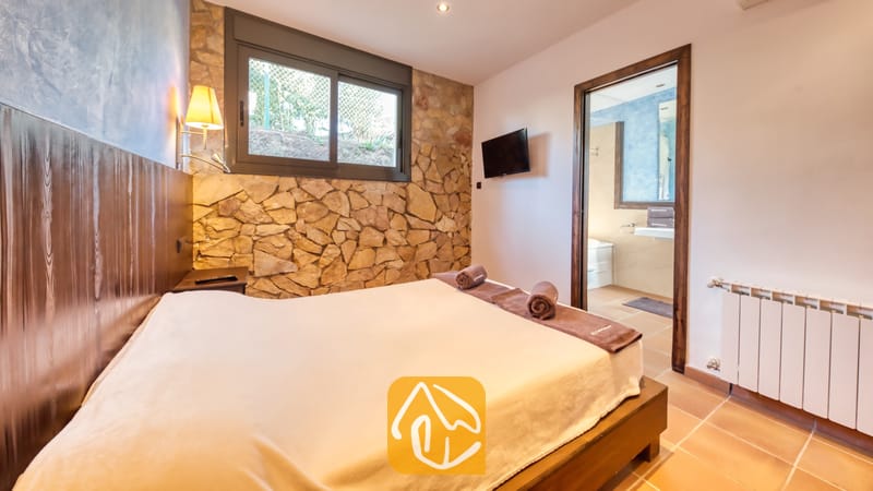 Villas de vacances Costa Brava Countryside Espagne - Villa Can Bernardi - Chambre avec un lit double, des serviettes marron enroulées, un mur d'accent en pierre et une porte ouverte menant à une salle de bains attenante.