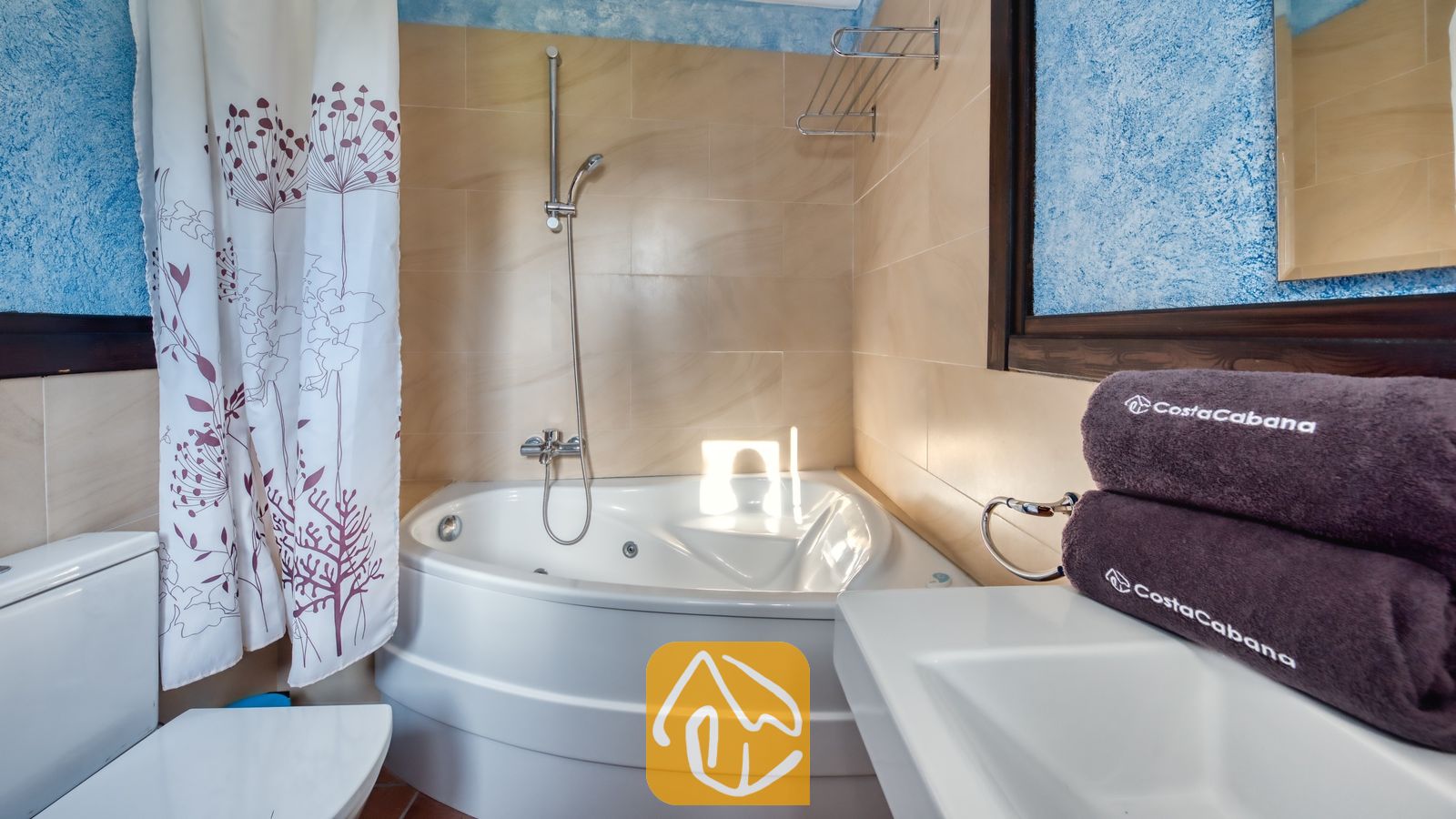 Vakantievilla's Costa Brava Countryside Spanje - Villa Can Bernardi - Hoekbad met chroomkleurige douchemranituren in een betegelde badkamer met blauwe, getextureerde muren en opgerolde bruine handdoeken op het wastafelblad.