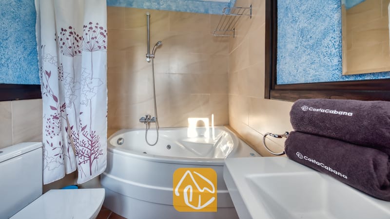 Villas de vacances Costa Brava Countryside Espagne - Villa Can Bernardi - Baignoire d'angle avec robinetterie de douche chromée dans une salle de bains carrelée aux murs texturés bleus, avec des serviettes marron enroulées sur le plan de toilette.