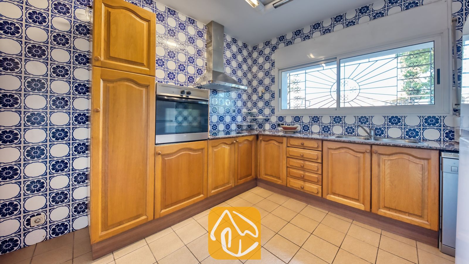 Vakantievilla's Costa Brava Countryside Spanje - Villa Can Bernardi - Keuken met houten kasten en blauw-gemzen wandtegels, voorzien van een ingebouwde oven, afzuigkap en een raam met rolluiken/veiligheidsroosters.