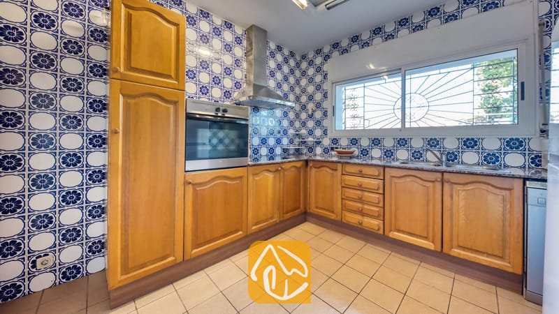 Villas de vacances Costa Brava Countryside Espagne - Villa Can Bernardi - Cuisine équipée de placards en bois et de carreaux muraux à motifs bleus, comprenant un four encastré, une hotte aspirante et une fenêtre pourvue de grilles de sécurité.