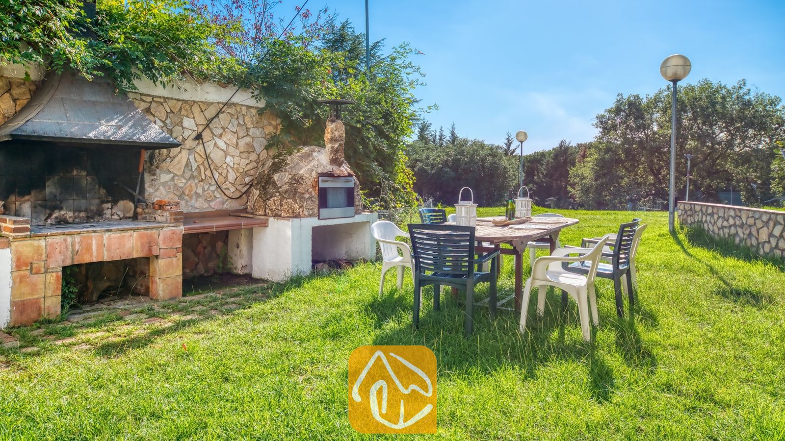 Vakantievilla's Costa Brava Countryside Spanje - Villa Can Bernardi - Buiten dineren op het gazon van Villa Can Bernardi, voorzien van een stenen barbecue, een houtoven, een houten eettafel en kunststof stoelen.