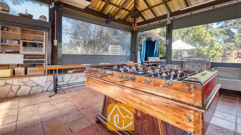 Vakantievilla's Costa Brava Countryside Spanje - Villa Can Bernardi - Houten tafelvoetbaltafel in een overdekt paviljoen bij Villa Can Bernardi, voorzien van tegelvloeren, geluidsapparatuur op planken en grote ramen met uitzicht op bomen.