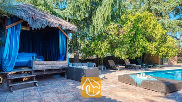 Vakantievilla's Costa Brava Countryside Spanje - Villa Can Bernardi - Rieten prieel met blauwe gordijnen en bank naast een zwembad met ligstoelen bij Villa Can Bernardi