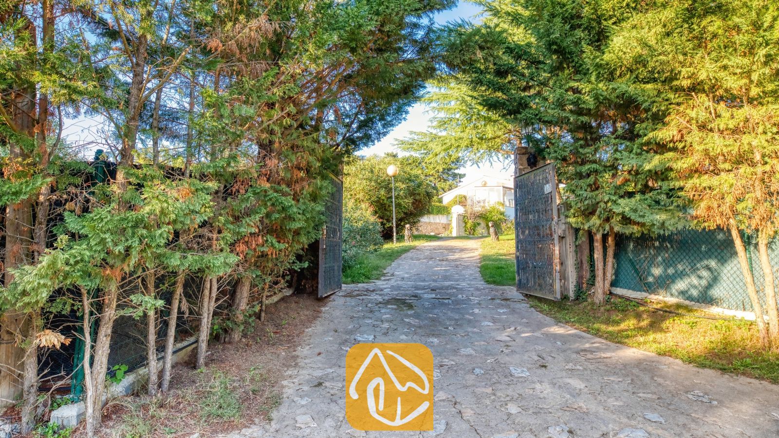 Vakantievilla's Costa Brava Countryside Spanje - Villa Can Bernardi - Een met stenen bestrate oprijlaan, omlijst door hoge groene bomen en openstaande metalen poorten, leidt naar de entree van Villa Can Bernardi