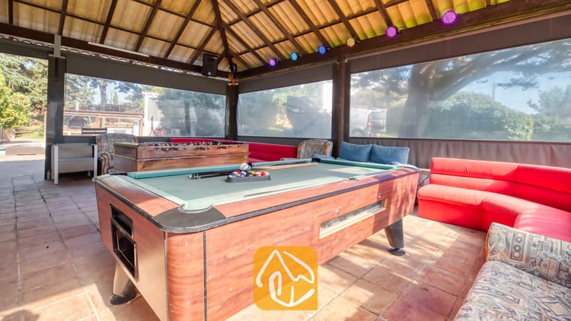 Vakantievilla's Costa Brava Countryside Spanje - Villa Can Bernardi - Overdekte buitenrecreatieruimte bij Villa Can Bernardi, voorzien van een houten biljarttafel, een tafelvoetbalspel en rode en gedessinieerde banken onder een pannendak.