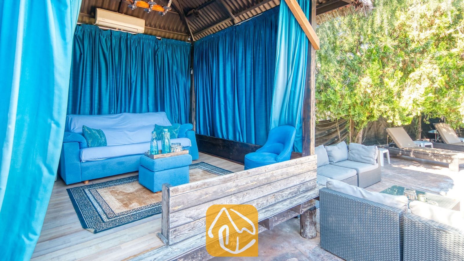 Vakantievilla's Costa Brava Countryside Spanje - Villa Can Bernardi - Overdekte buitenlounge bij Villa Can Bernardi, voorzien van helderblauwe gordijnen, een bijpassende blauwe bank en een vloerkleed met patroon.