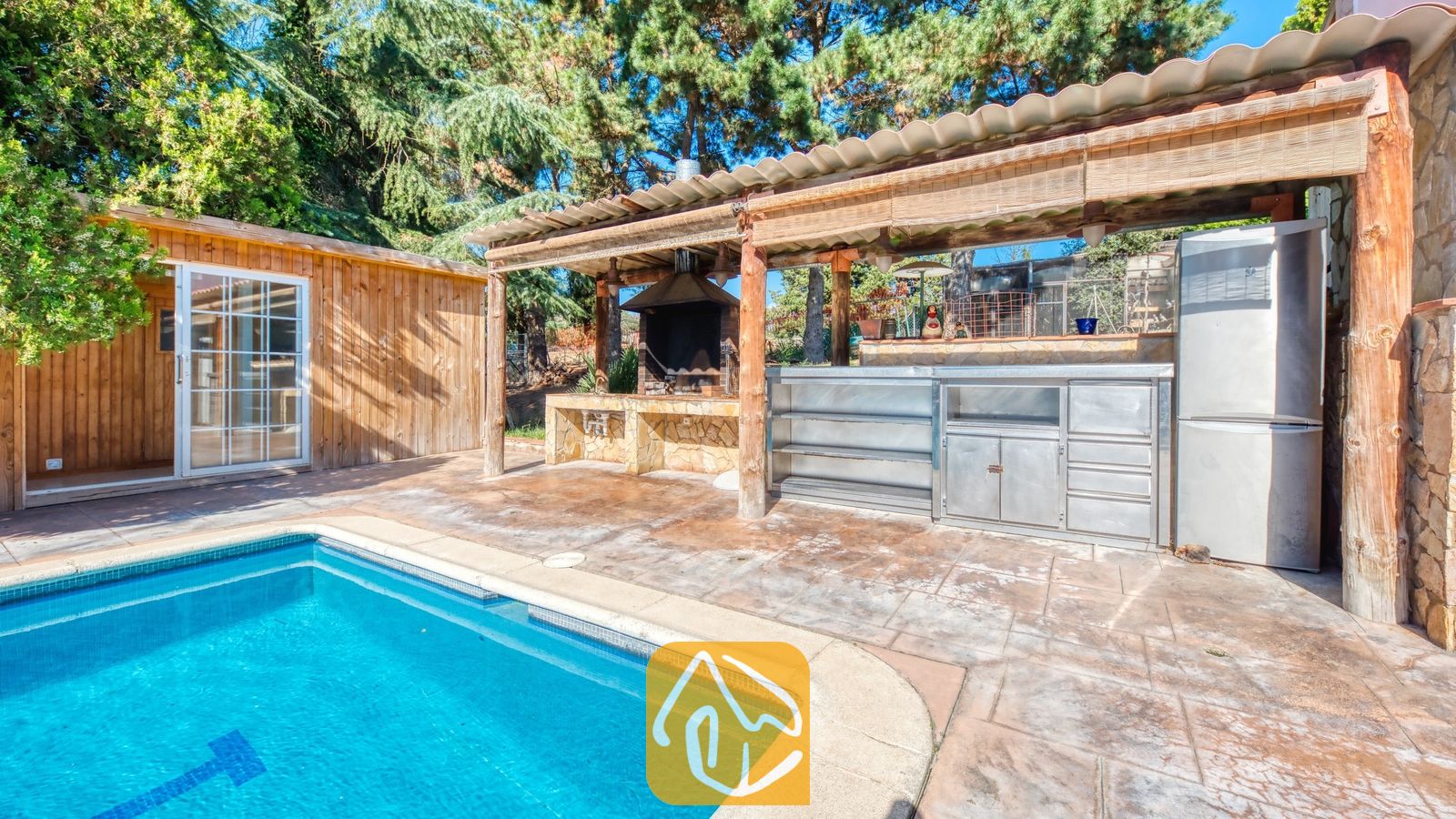 Vakantievilla's Costa Brava Countryside Spanje - Villa Can Bernardi - Buitenkeuken met barbecue, een metalen aanrecht en koelkast onder een houten pergola naast het zwembad van Villa Can Bernardi