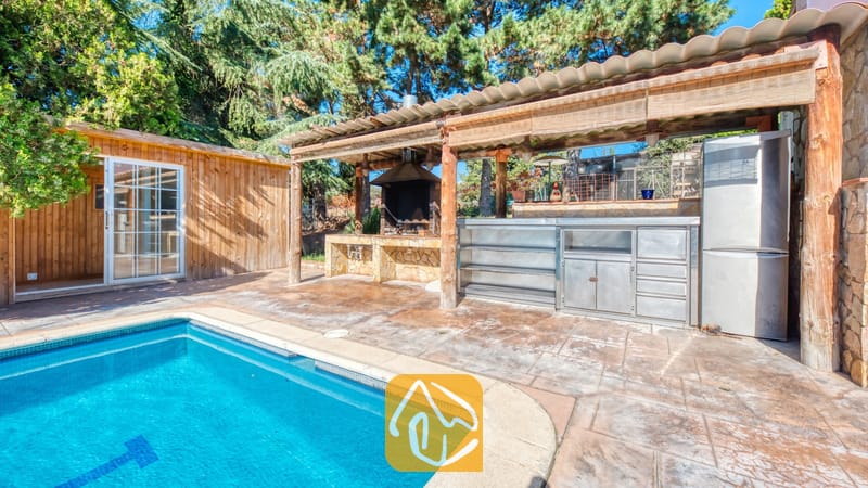 Vakantievilla's Costa Brava Countryside Spanje - Villa Can Bernardi - Buitenkeuken met barbecue, een metalen aanrecht en koelkast onder een houten pergola naast het zwembad van Villa Can Bernardi