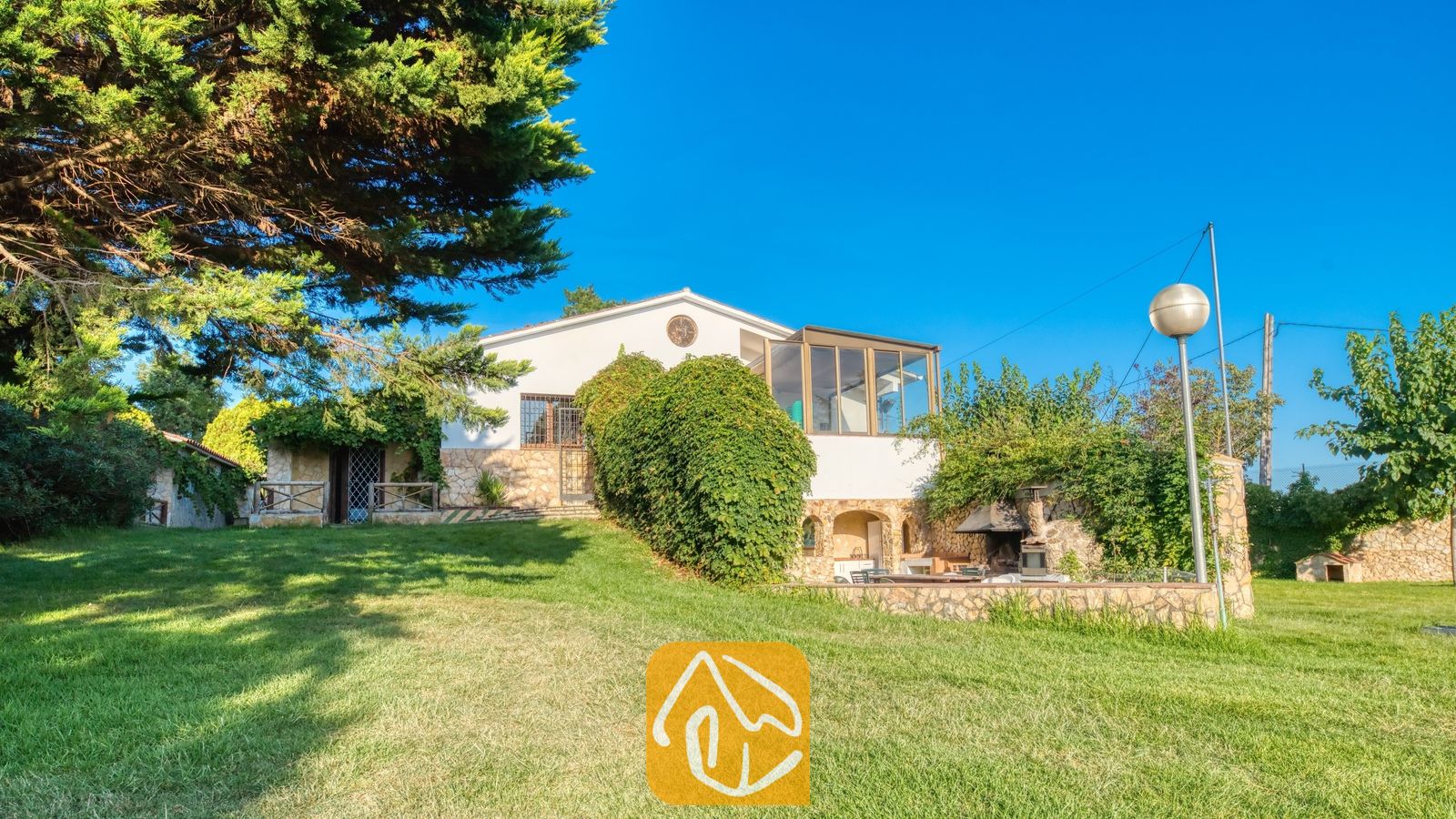 Vakantievilla's Costa Brava Countryside Spanje - Villa Can Bernardi - Buitenaanzicht van Villa Can Bernardi met een witte gevel, stenen sokkelmuren, groene gazons en een buitenbarbecue onder een helderblauwe hemel