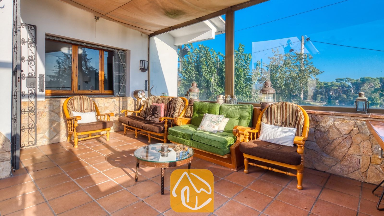 Vakantievilla's Costa Brava Countryside Spanje - Villa Can Bernardi - Overdekt terras bij Villa Can Bernardi, voorzien van bruine en groene houten fauteuils, een salontafel met glazen blad, stenen muren en grote glazen ramen met uitzicht op de bomen.