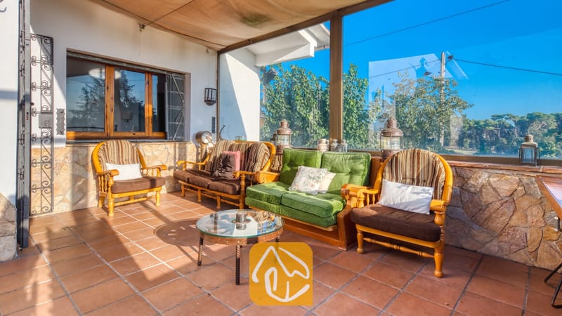 Vakantievilla's Costa Brava Countryside Spanje - Villa Can Bernardi - Overdekt terras bij Villa Can Bernardi, voorzien van bruine en groene houten fauteuils, een salontafel met glazen blad, stenen muren en grote glazen ramen met uitzicht op de bomen.