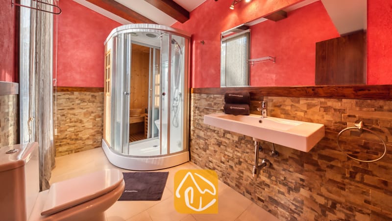 Villas de vacances Costa Brava Countryside Espagne - Villa Can Bernardi - Salle de bain dotée d'une cabine de douche incurvée en verre avec un intérieur en bois de sauna, d'un lavabo blanc suspendu et de murs carrelés en pierre et de couleur rouge.