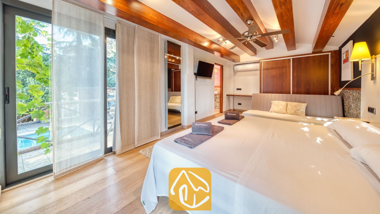 Vakantievilla's Costa Brava Countryside Spanje - Villa Can Bernardi - Slaapkamer in Villa Can Bernardi, voorzien van een groot bed met opgevouwen handdoeken, houten plafondbalken en een glazen schuifdeur die uitzicht biedt op het zwembad.
