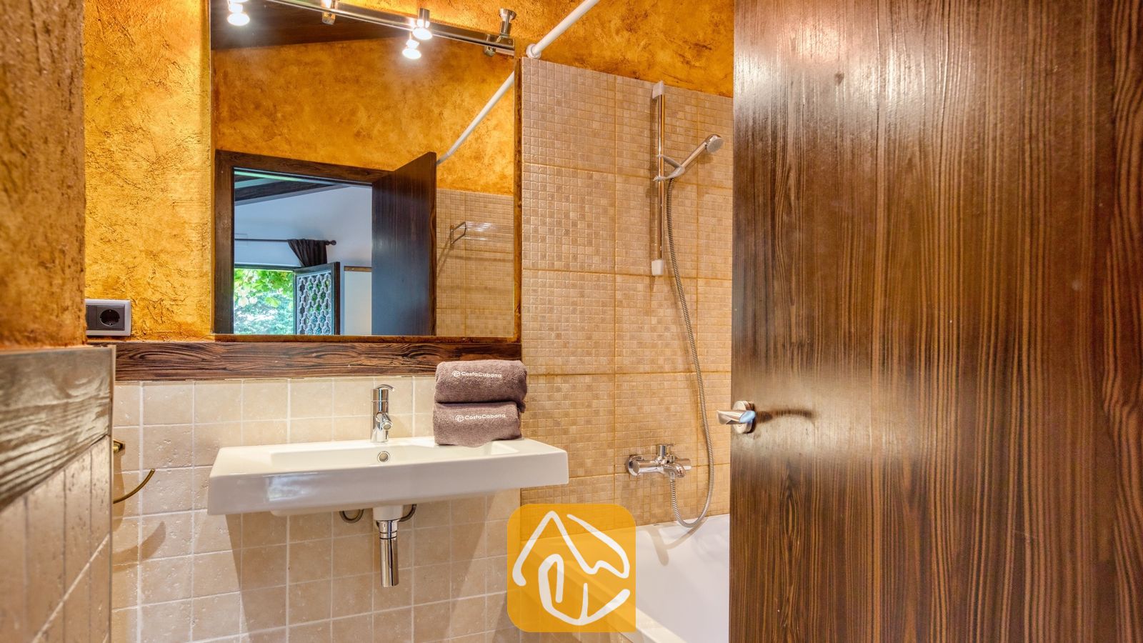Vakantievilla's Costa Brava Countryside Spanje - Villa Can Bernardi - Badkamer met een witte wandwastafel, betegelde douche boven bad en opgevouwen bruine handdoeken weerspiegeld in de grote badkamerspiegel.