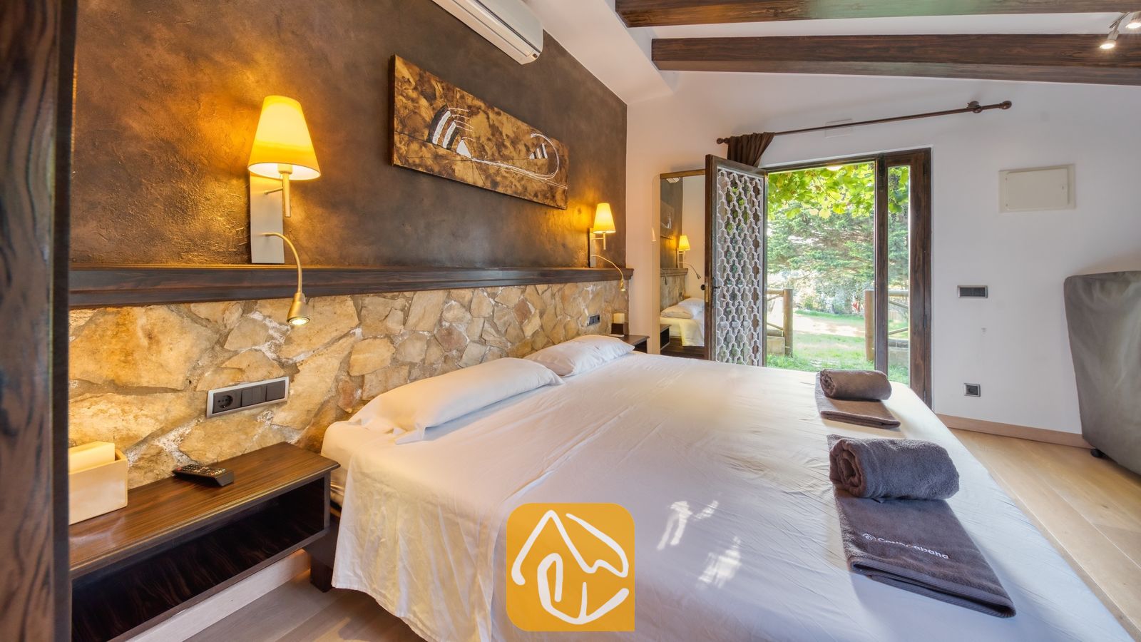 Vakantievilla's Costa Brava Countryside Spanje - Villa Can Bernardi - Slaapkamer met tweepersoonsbed, stenen hoofdbord, donkerhouten nachtkastjes en open glazen deuren die uitkomen op het groen bij Villa Can Bernardi
