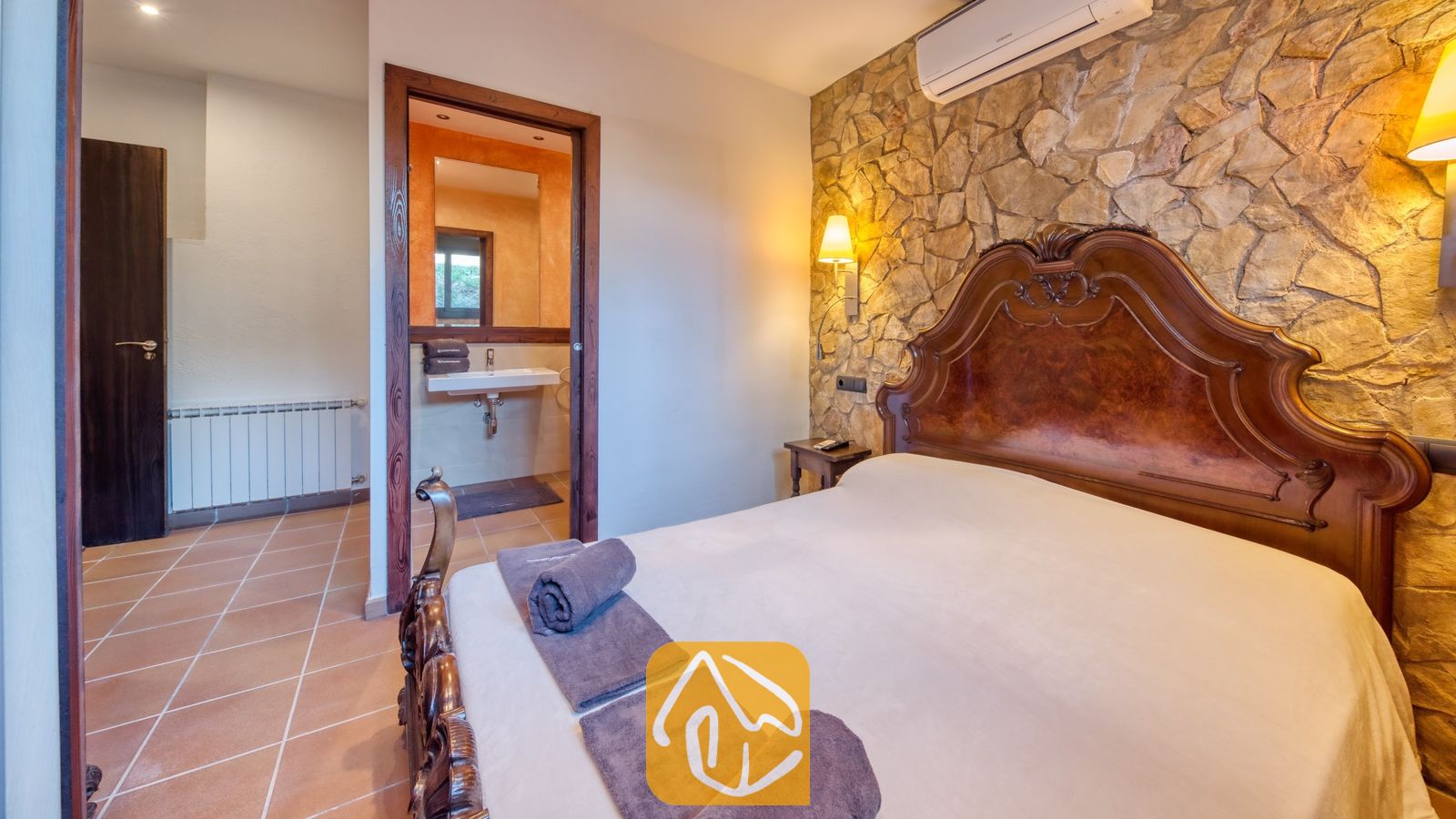 Vakantievilla's Costa Brava Countryside Spanje - Villa Can Bernardi - Slaapkamer met een sierlijk houten hoofdbord tegen een stenen accentmuur, opgerolde grijze handdoeken op het bed en uitzicht op de en-suite badkamer door de open deuropening.