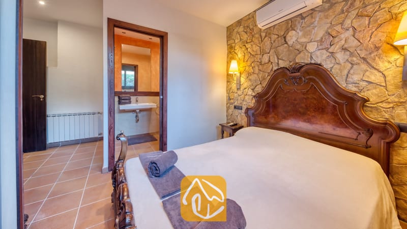Villas de vacances Costa Brava Countryside Espagne - Villa Can Bernardi - Chambre dotée d'une tête de lit en bois ciselé adossée à un mur d'accent en pierre, avec des serviettes grises roulées sur le lit et un aperçu de la salle de bains attenante à travers la porte ouverte.