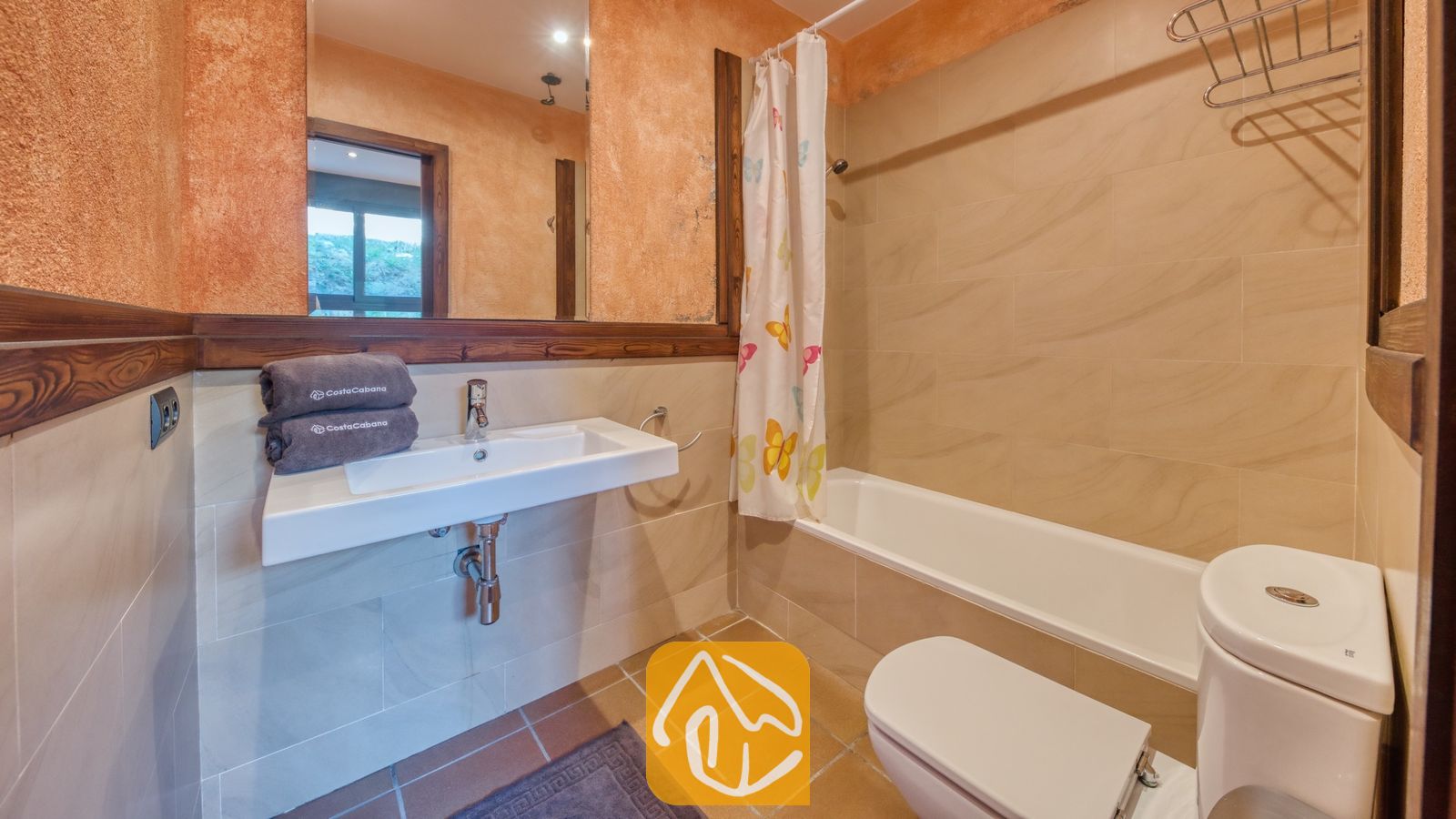 Vakantievilla's Costa Brava Countryside Spanje - Villa Can Bernardi - Badkamer voorzien van een witte wastafel met opgevouwen grijze handdoeken, een ligbad met een vlinderdouchegordijn, een toilet en getextureerde terracotta muren.