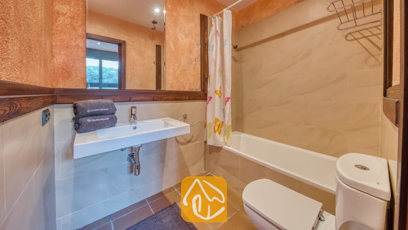 Villas de vacances Costa Brava Countryside Espagne - Villa Can Bernardi - Salle de bains comprenant un lavabo blanc avec des serviettes grises pliées, une baignoire dotée d'un rideau de douche à motif de papillons, des toilettes et des murs en terracotta texturée.