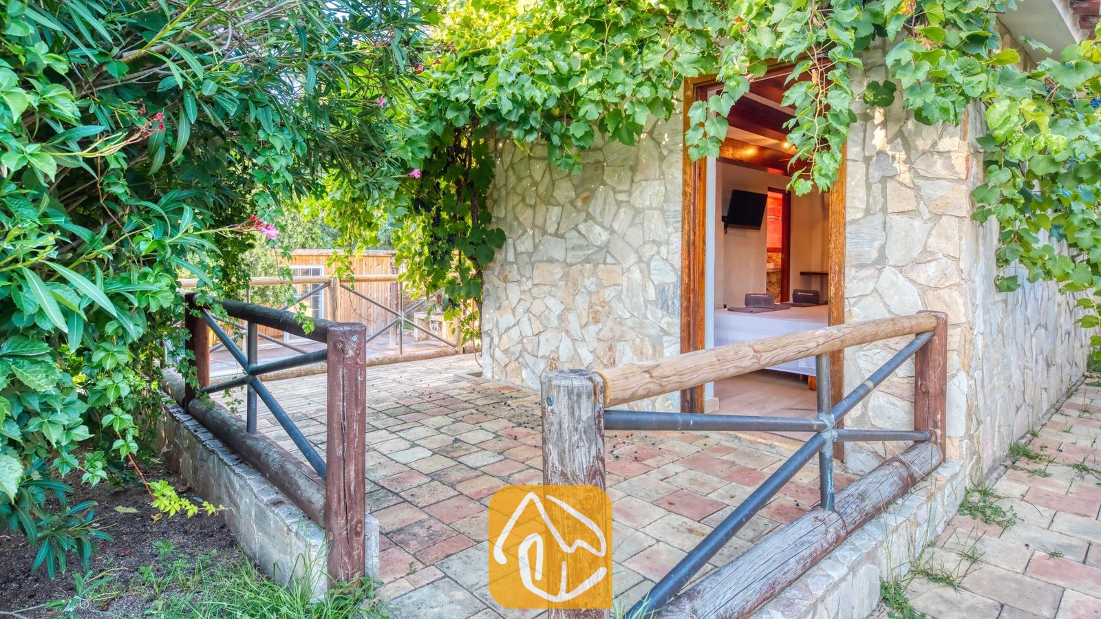 Vakantievilla's Costa Brava Countryside Spanje - Villa Can Bernardi - Stenen buitenkant van Villa Can Bernardi met een houten balkon en balustrade, omringd door weelderige groene klimplanten en gebladerte dat leidt naar een open deuropening.