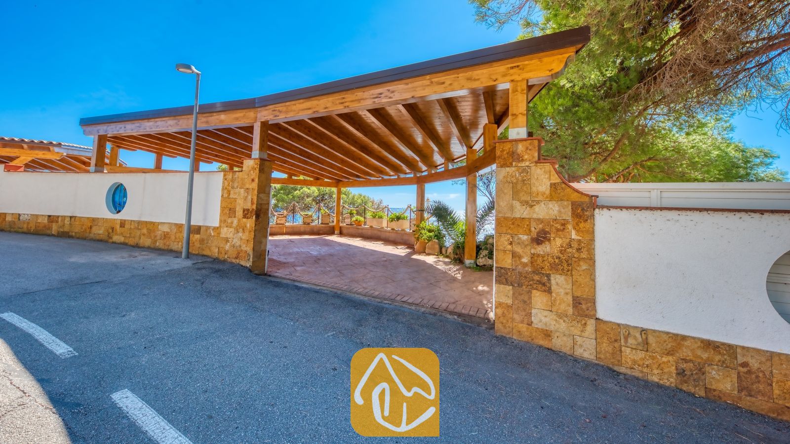 Vakantievilla's Costa Brava Spanje - Villa Infinity - Overdekte houten pergola-ingang van Villa Infinity, gekenmerkt door stenen pilaren en witte muren rondom, met een asfaloweg op de voorgrond onder een helderblauwe hemel.
