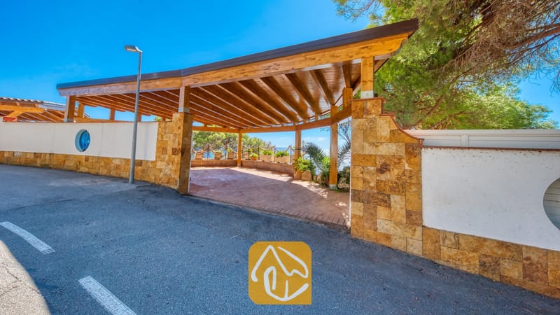 Villas de vacances Costa Brava Espagne - Villa Infinity - Entrée sous une pergola en bois couverte à la Villa Infinity, agrémentée de piliers en pierre et de murs d'enceinte blancs, avec une route asphaltée au premier plan sous un ciel d'un bleu éclatant.