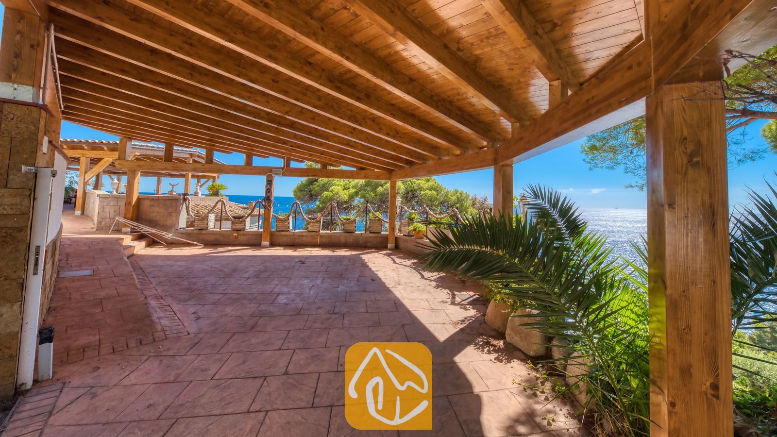 Vakantievilla's Costa Brava Spanje - Villa Infinity - Overdekte houten pergola bij Villa Infinity met betegelde stenen vloer, uitkijkend over de zee en groene bomen onder een helderblauwe hemel.