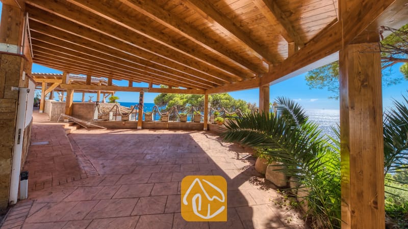 Villas de vacances Costa Brava Espagne - Villa Infinity - Pergola en bois couverte à la Villa Infinity avec sol en pierre carrelée, surplombant la mer et les arbres verdoyants sous un ciel d'un bleu éclatant.