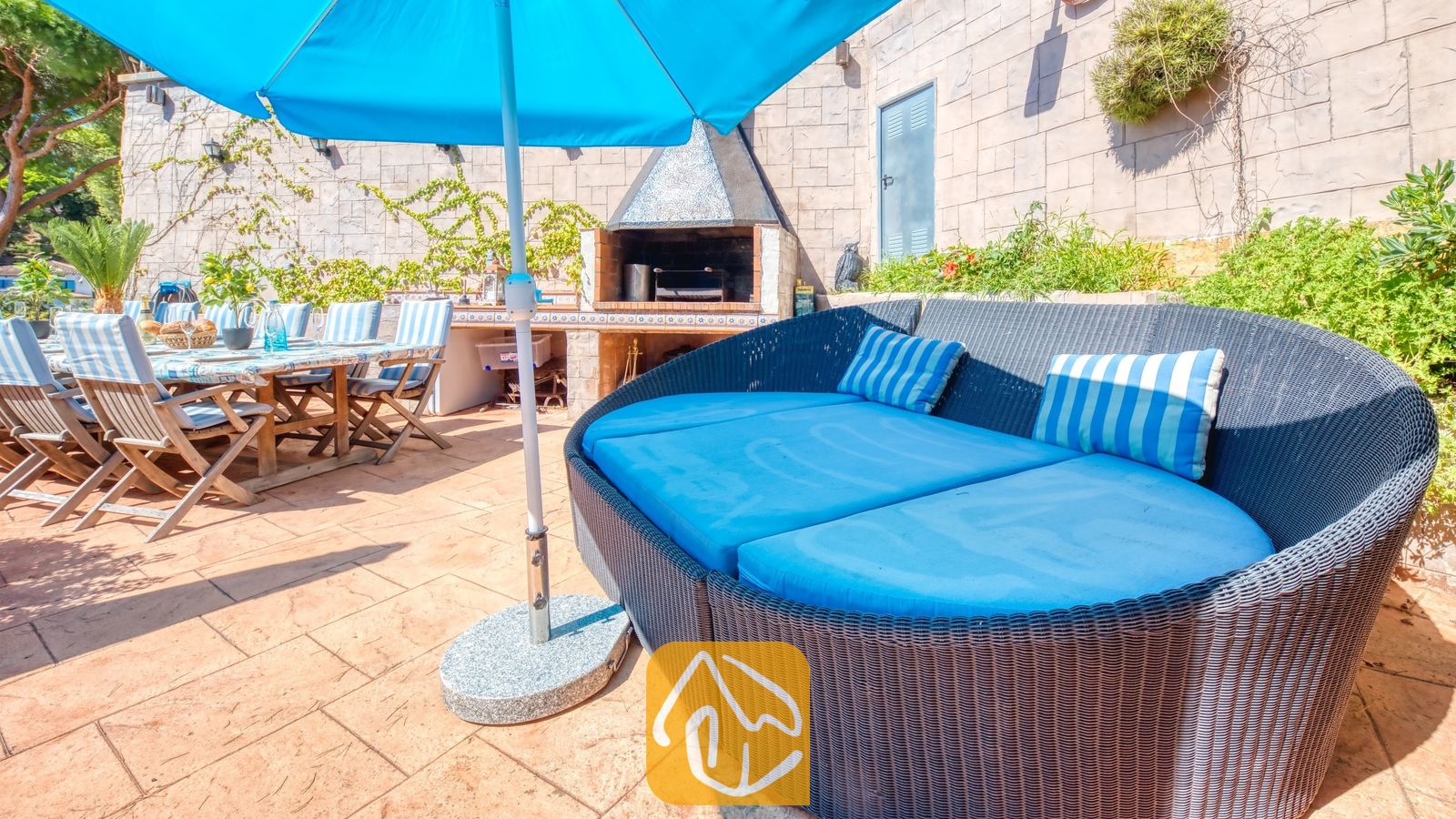 Vakantievilla's Costa Brava Spanje - Villa Infinity - Gebogen buitenbank van rotan met helderblauwe kussens en gestreepte sierkussens op een stenen terras bij Villa Infinity, met een stenen barbecue en eettafel op de achtergrond.